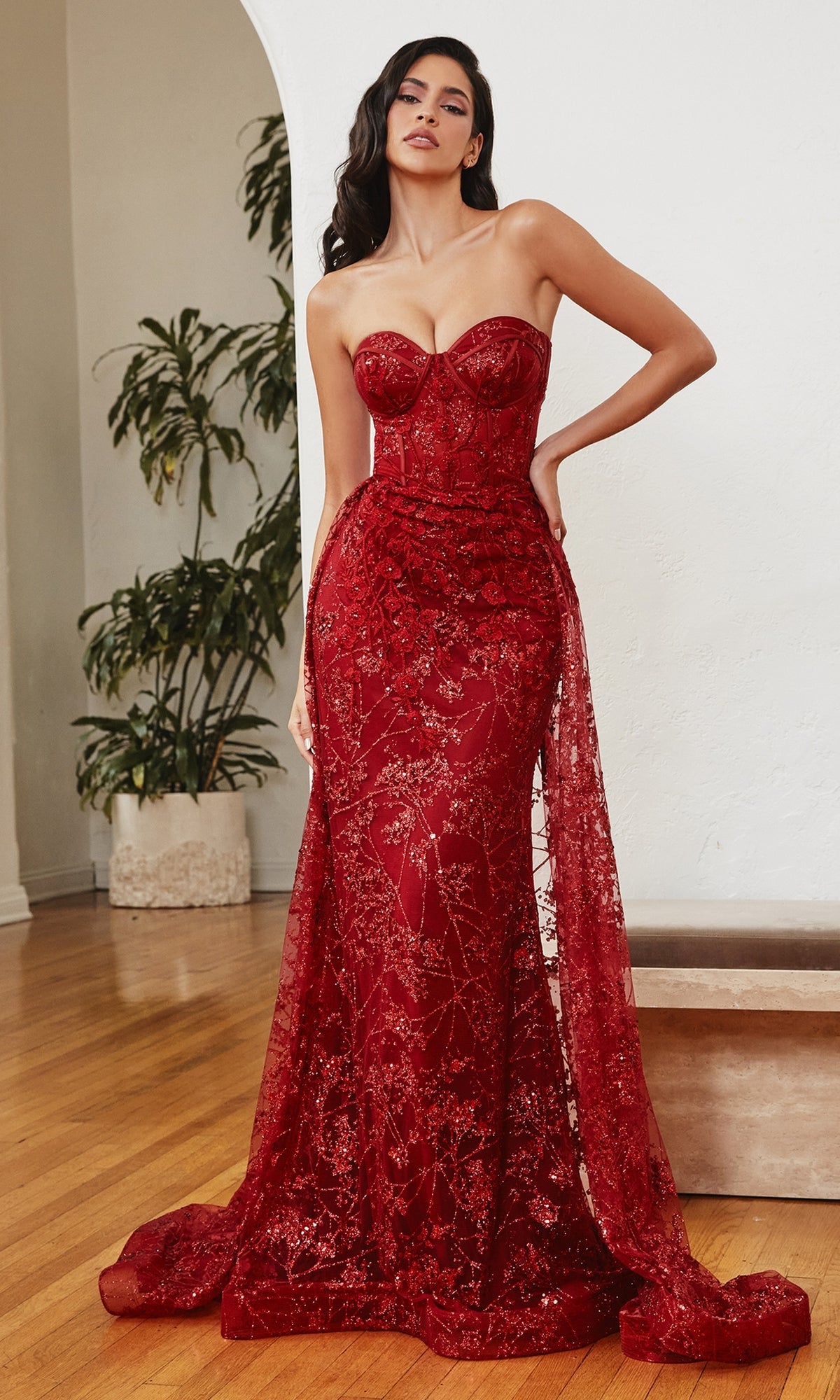 A woman stands indoors on a wooden floor by a potted plant, wearing the Glitter-Print Strapless Corset Prom Dress CB046—a fitted long red evening gown with a sheer overlay and sequin details.
